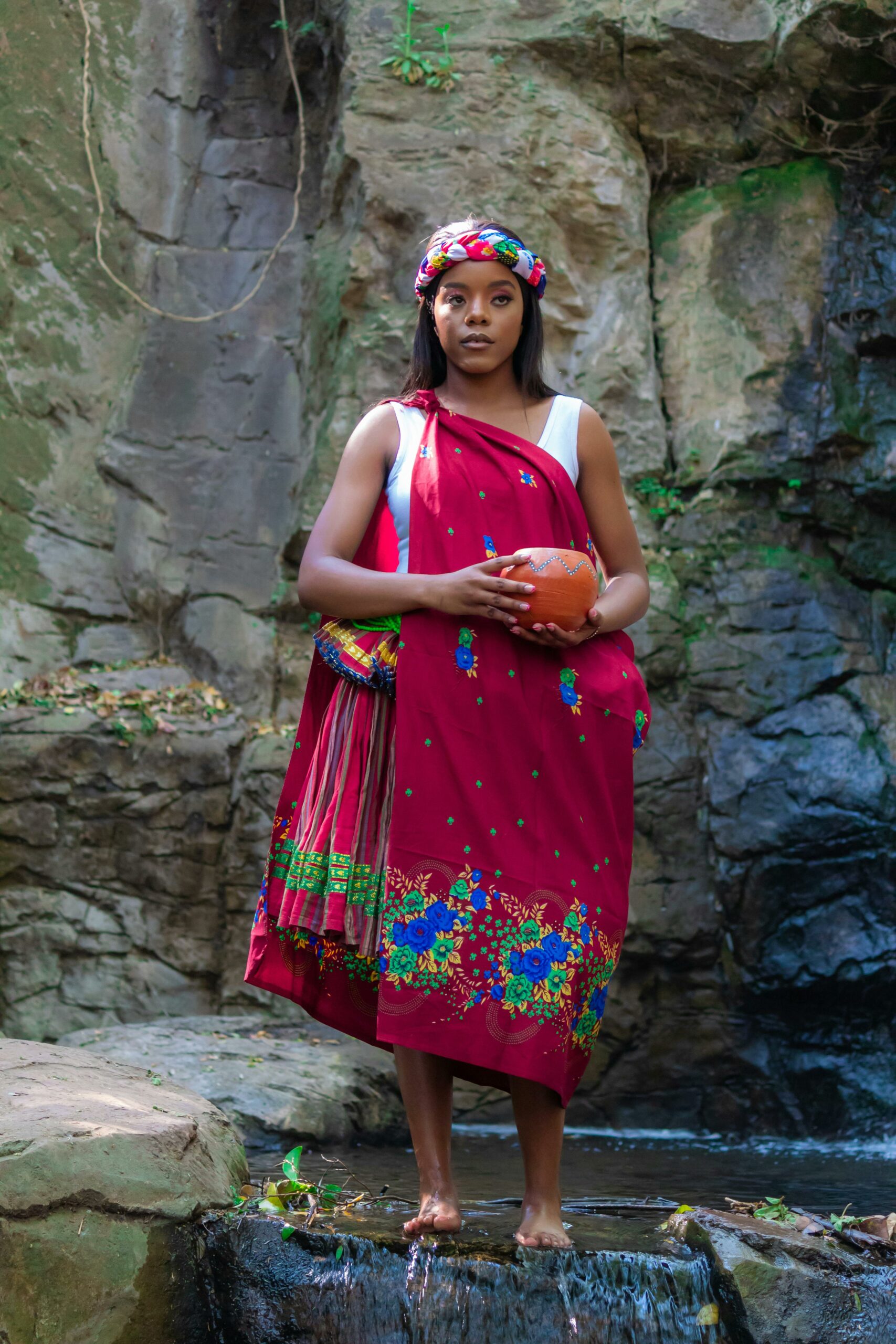 Woman posing in vibrant traditional dress near a rocky waterfall showcasing natural beauty.
